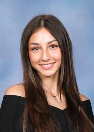 Young woman with long dark hair wearing an off-shoulder black top, smiling at camera against a blue background