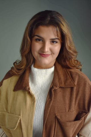 Young woman with shoulder-length reddish-brown hair wearing a tan corduroy jacket over a white ribbed sweater, smiling at the camera against a gray background