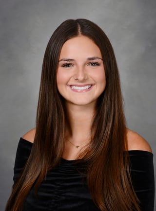 Portrait of a young woman with long brown hair wearing a black off-shoulder top, smiling at the camera against a gray background