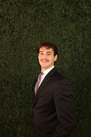 Professional headshot of a smiling man in a black blazer and purple dress shirt against a green textured background