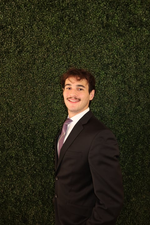 Professional headshot of a smiling man in a black blazer and purple dress shirt against a green textured background