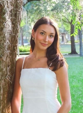 Woman in white sleeveless top posing against tree in park with green grass and trees in background