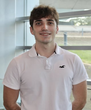 Young man wearing a white polo shirt smiling at the camera with large windows and outdoor field visible behind him